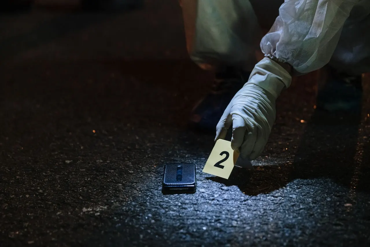 A gloved crime scene investigator places a numbered evidence marker beside a phone on night pavement, the scene work California bachelor's degrees in forensic science prepare for