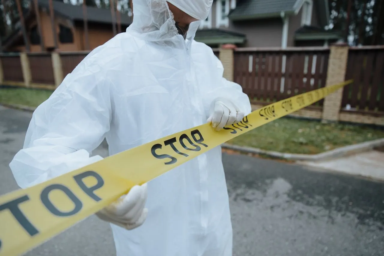 An investigator in white coveralls and gloves lifts a length of yellow barrier tape across a suburban street, the scene-control work behind online crime scene investigation bachelor's degrees for California students