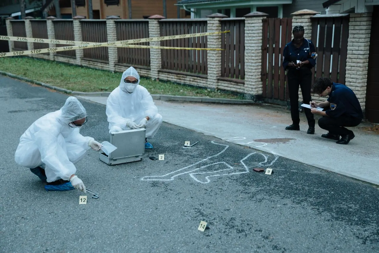 Two technicians in white coveralls photograph and measure evidence markers around a chalk body outline while officers take notes behind the tape, the documented procedure California forensic science degree requirements train students to follow
