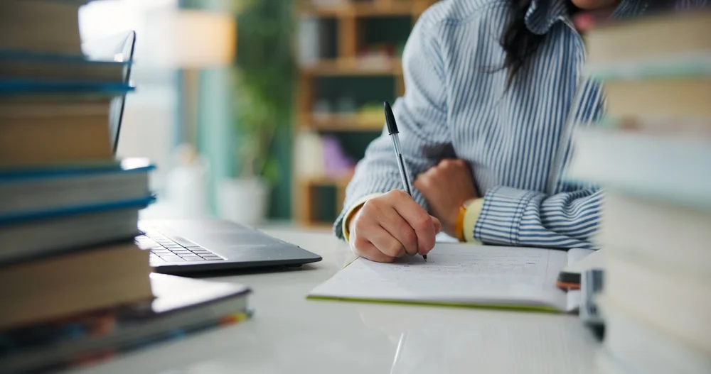 Close-up of a student writing beside a stack of books